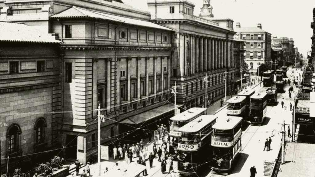 Historic black-and-white photo of a busy city street with several double-decker trams, people walking, and large classical buildings lining the road—like visual cover versions of vibrant urban life from another era.