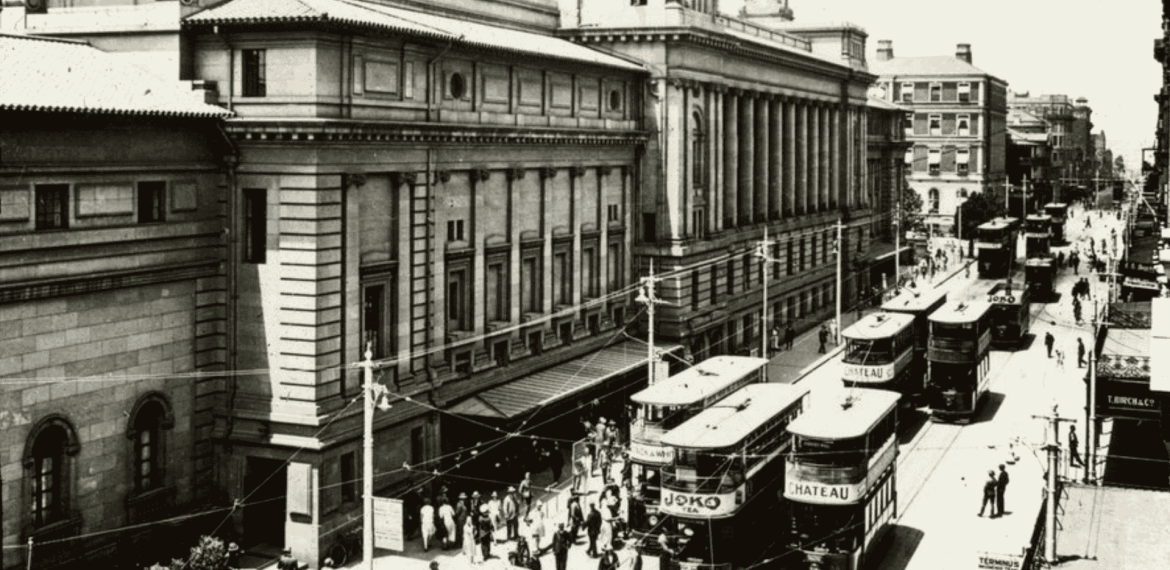 Historic black-and-white photo of a busy city street with several double-decker trams, people walking, and large classical buildings lining the road—like visual cover versions of vibrant urban life from another era.