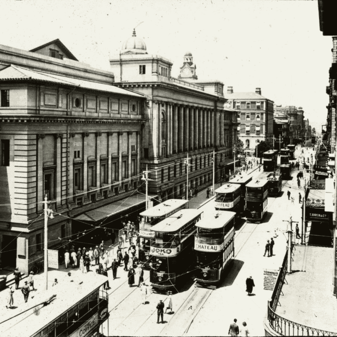 Black-and-white photo of a busy city street with several double-decker trams, pedestrians on sidewalks, and a large classical building with columns and domes—an early 20th-century urban scene reminiscent of cover versions in architecture.
