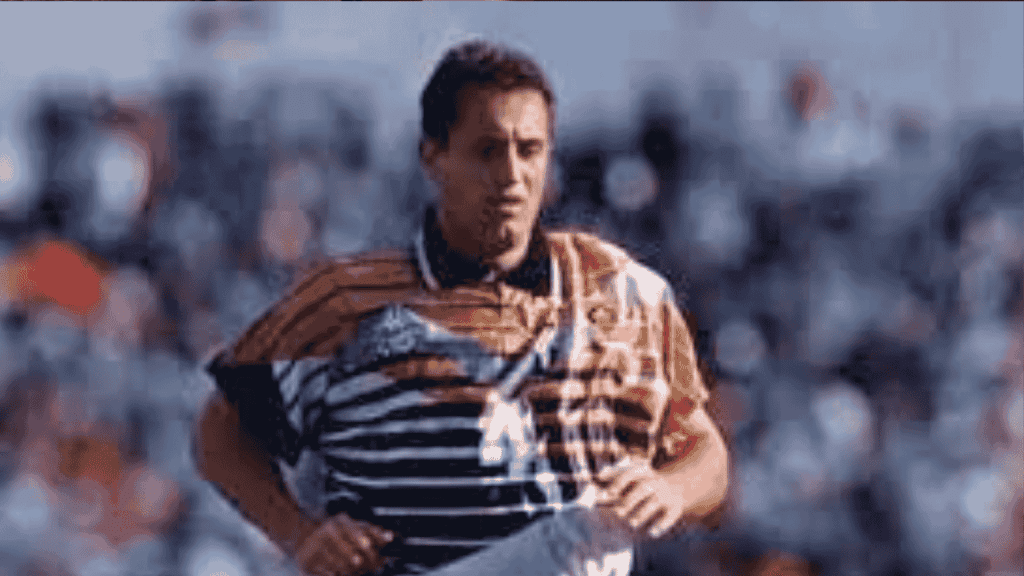 Mark McNulty, in a distinctive orange, black, and white striped jersey, is running on the field as blurred spectators watch from the background.