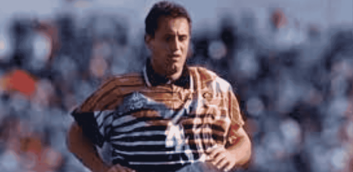 Mark McNulty, in a distinctive orange, black, and white striped jersey, is running on the field as blurred spectators watch from the background.