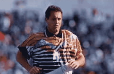 Mark McNulty, in a distinctive orange, black, and white striped jersey, is running on the field as blurred spectators watch from the background.