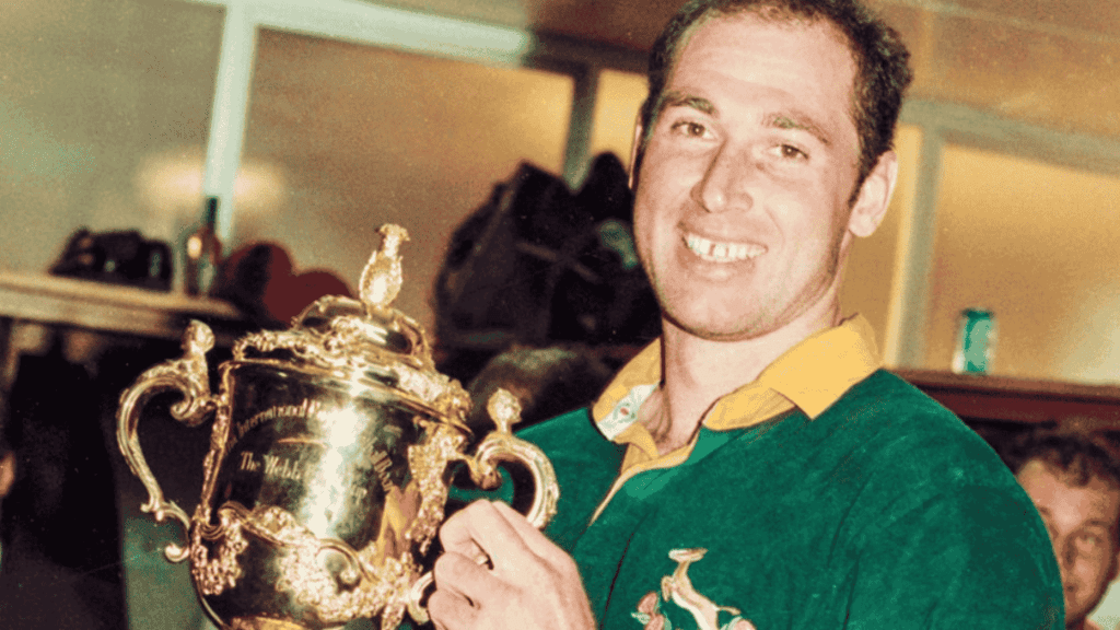 A smiling rugby player in a green jersey holds up a gold trophy cup, celebrating a cross-border victory indoors.