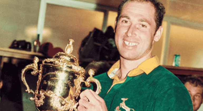 A smiling rugby player in a green jersey holds up a gold trophy cup, celebrating a cross-border victory indoors.