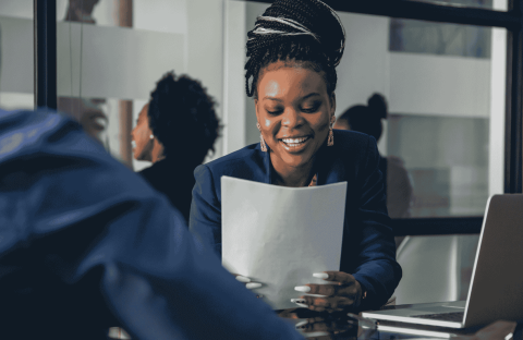 A woman smiling while holding papers sits at a desk in a modern office, engaging in conversation about business opportunities with another person whose back is visible in the foreground.