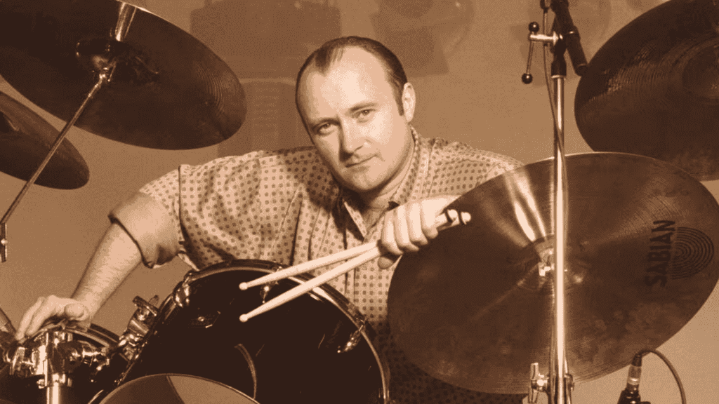 A man with a receding hairline and patterned shirt sits at a drum set, holding drumsticks and leaning on a Sabian cymbal, looking directly at the camera in a sepia tone—reminiscent of Phil Collins.