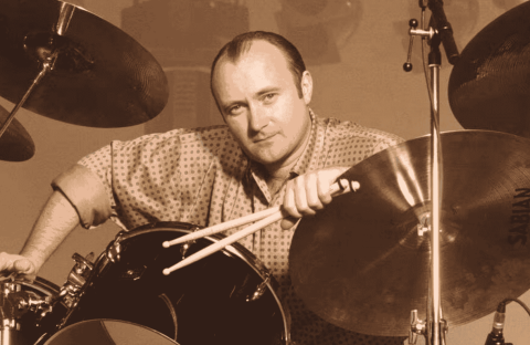 A man with a receding hairline and patterned shirt sits at a drum set, holding drumsticks and leaning on a Sabian cymbal, looking directly at the camera in a sepia tone—reminiscent of Phil Collins.