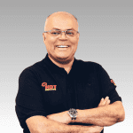A smiling man with glasses wears a plain black polo shirt with the HOT 102FM logo, standing with arms crossed against a gray gradient background.