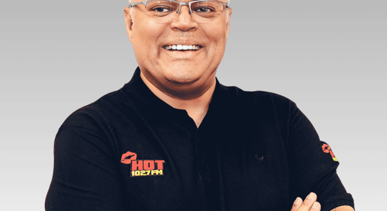 A smiling man with glasses wears a plain black polo shirt with the HOT 102FM logo, standing with arms crossed against a gray gradient background.