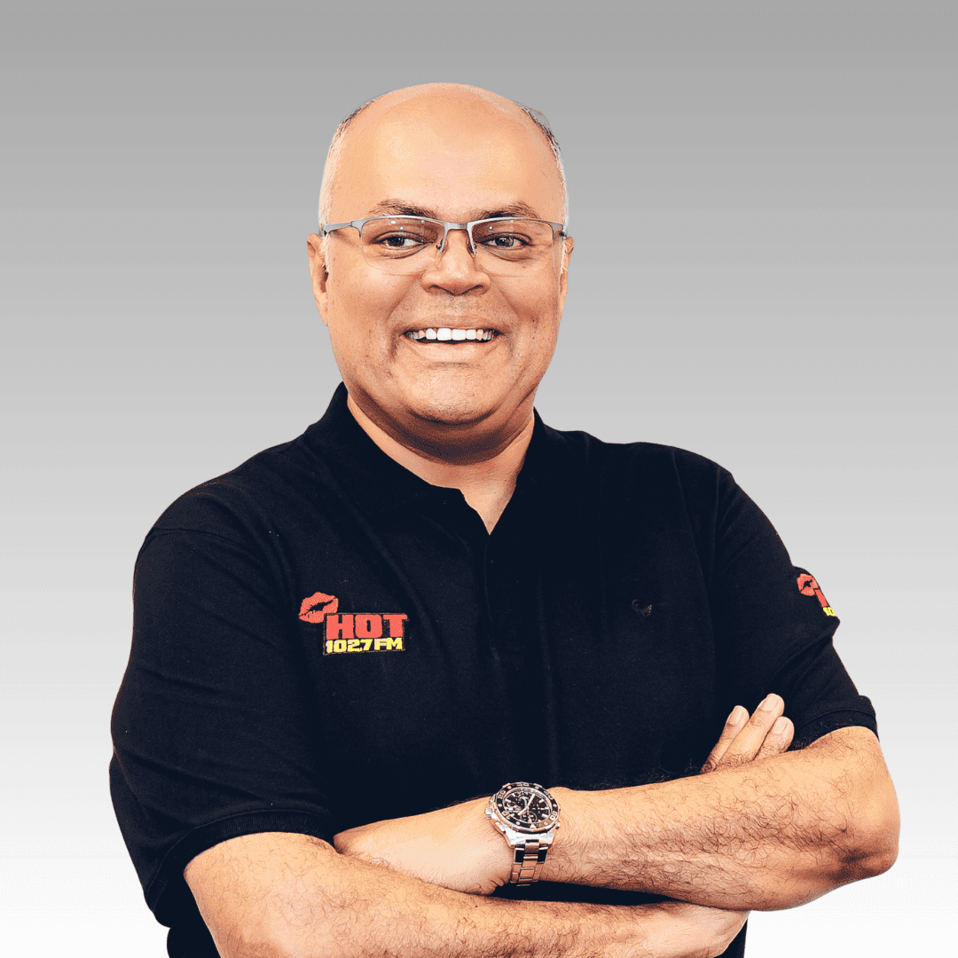 A smiling man with glasses wears a plain black polo shirt with the HOT 102FM logo, standing with arms crossed against a gray gradient background.