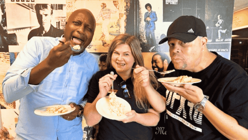 Three people smiling and eating slices of cake with forks, standing in front of a wall covered with various vintage music and film posters—celebrating their Matric achievements together.