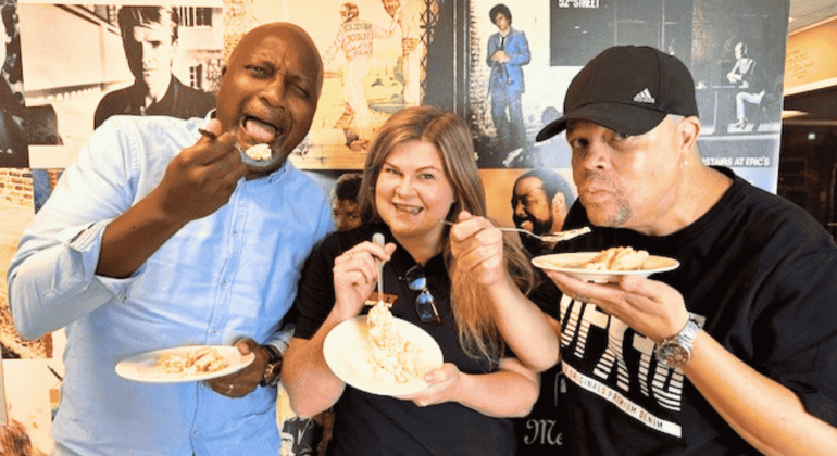 Three people smiling and eating slices of cake with forks, standing in front of a wall covered with various vintage music and film posters—celebrating their Matric achievements together.