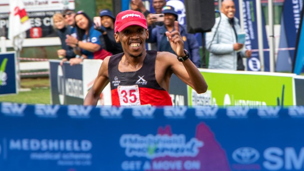 A male runner wearing a red cap and black-red outfit, bib number 35, smiles and raises his hand in celebration as he crosses the finish line at a race, just like he did last Christmas, with cheering spectators and banners in the background.