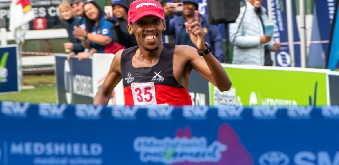 A male runner wearing a red cap and black-red outfit, bib number 35, smiles and raises his hand in celebration as he crosses the finish line at a race, just like he did last Christmas, with cheering spectators and banners in the background.