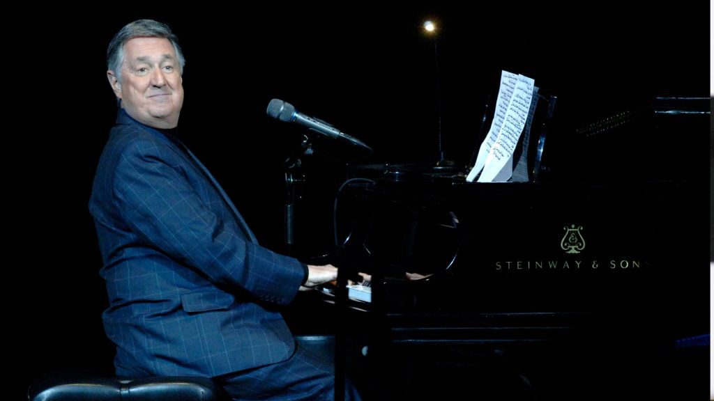 A man in a dark suit sits at a Steinway & Sons grand piano on stage, smiling at the audience. Sheet music is on the piano, and a microphone hovers above the keys as he prepares to perform a Blancmange classic. The background is dark.
