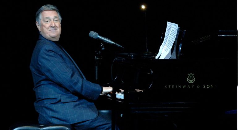 A man in a dark suit sits at a Steinway & Sons grand piano on stage, smiling at the audience. Sheet music is on the piano, and a microphone hovers above the keys as he prepares to perform a Blancmange classic. The background is dark.