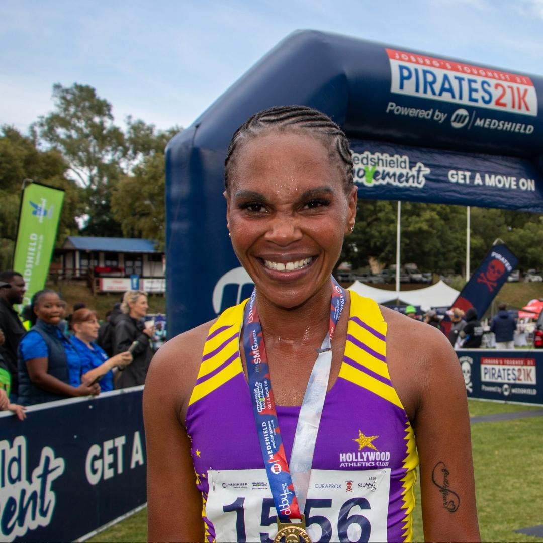 A smiling runner wearing a purple and yellow athletic jersey and a medal stands in front of a race finish line arch at an outdoor event, reliving the thrill of last Christmas, with other people and banners visible in the background.