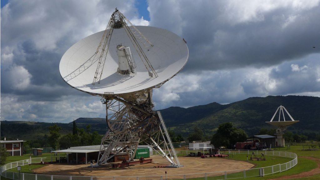 A large white radio telescope dish pointed toward the sky, set in a fenced area with green hills and trees in the background. Another smaller dish is visible farther away under a partly cloudy sky, evoking the legacy of Miriam Makeba reaching out to new horizons.