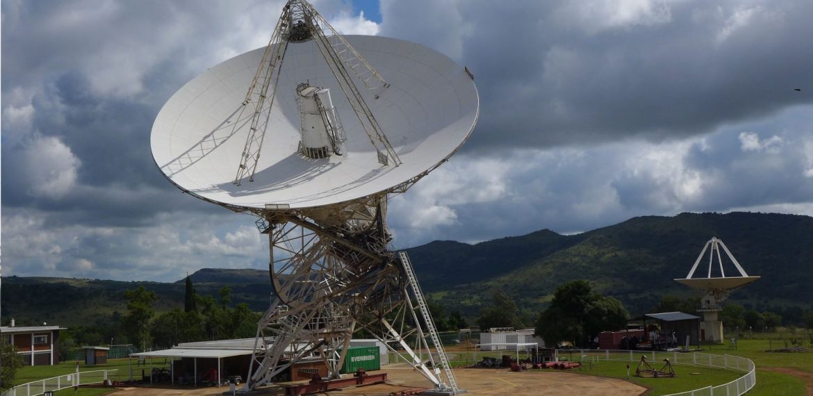 A large white radio telescope dish pointed toward the sky, set in a fenced area with green hills and trees in the background. Another smaller dish is visible farther away under a partly cloudy sky, evoking the legacy of Miriam Makeba reaching out to new horizons.