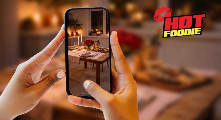 A person holds a phone, capturing a Valentine's dinner table adorned with candles and flowers. The background is blurred. “HOT FOODIE” with a red lips graphic is displayed in the upper right corner.
