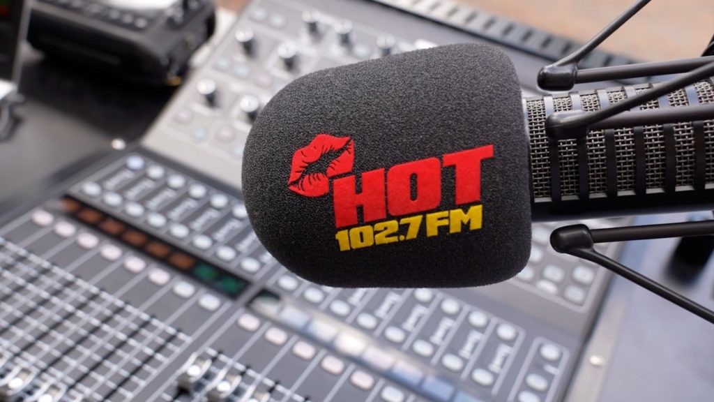 A microphone with a black foam cover displaying a red lipstick mark and the words HOT 102.7 FM in red and yellow, inspired by Miriam Makeba, positioned in front of a blurred audio mixing console.