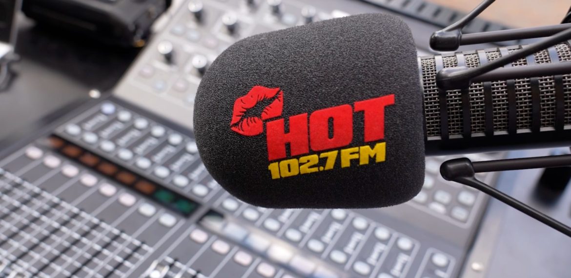 A microphone with a black foam cover displaying a red lipstick mark and the words HOT 102.7 FM in red and yellow, inspired by Miriam Makeba, positioned in front of a blurred audio mixing console.