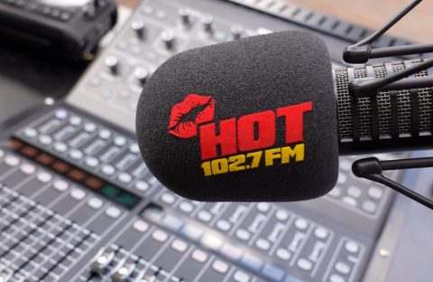 A microphone with a black foam cover displaying a red lipstick mark and the words HOT 102.7 FM in red and yellow, inspired by Miriam Makeba, positioned in front of a blurred audio mixing console.