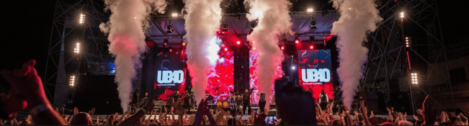 A concert stage with colorful lights, smoke effects, and the word UB40 displayed on large screens. The audience raises their hands, enjoying the live performance at night—a vibrant tribute echoing sounds reminiscent of Miriam Makeba.