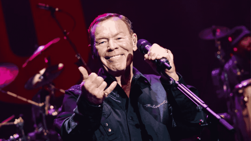 A smiling male singer holds a microphone and gestures with his hand on stage under purple lighting, performing in front of drums and other musicians—a vibrant scene reminiscent of Miriam Makeba's spirited concerts.