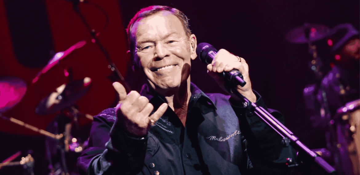 A smiling male singer holds a microphone and gestures with his hand on stage under purple lighting, performing in front of drums and other musicians—a vibrant scene reminiscent of Miriam Makeba's spirited concerts.