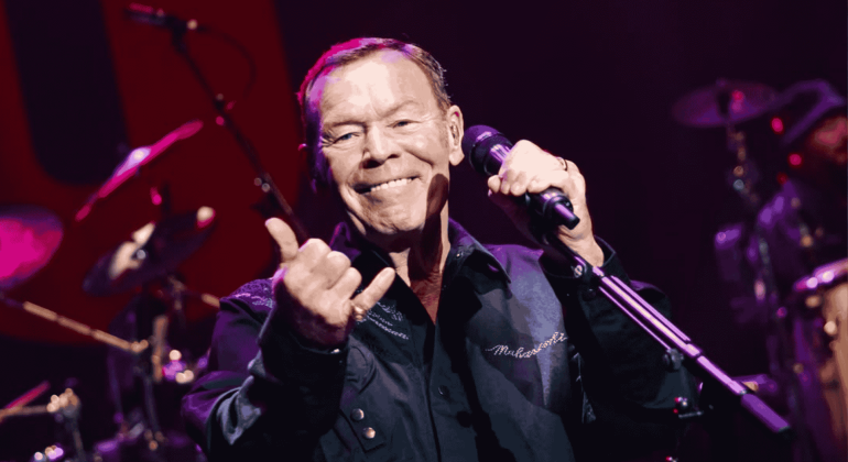 A smiling male singer holds a microphone and gestures with his hand on stage under purple lighting, performing in front of drums and other musicians—a vibrant scene reminiscent of Miriam Makeba's spirited concerts.