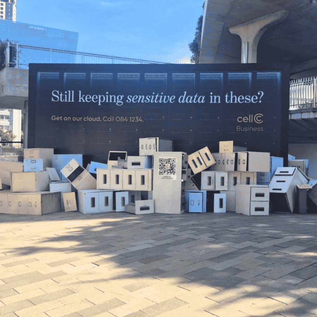 A large billboard reads, Still keeping sensitive data in these? above a pile of cardboard boxes and Matric files, promoting Cell C Business cloud services. Sunlight casts shadows on the pavement in front.