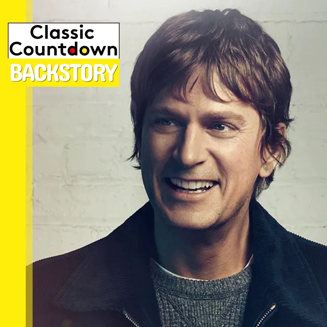 A smiling man with short brown hair wears a black jacket over a gray sweater, standing in front of a white brick wall. The text Classic Countdown BACKSTORY and “Last Christmas” appears in the top left corner on a yellow background.