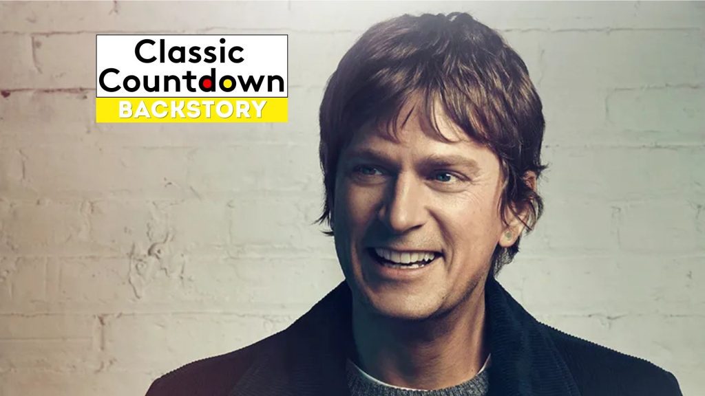 A smiling man with brown hair and a dark jacket stands in front of a light brick wall. In the upper left corner, a Classic Countdown Backstory logo appears, featuring bold black and yellow text inspired by "Last Christmas.