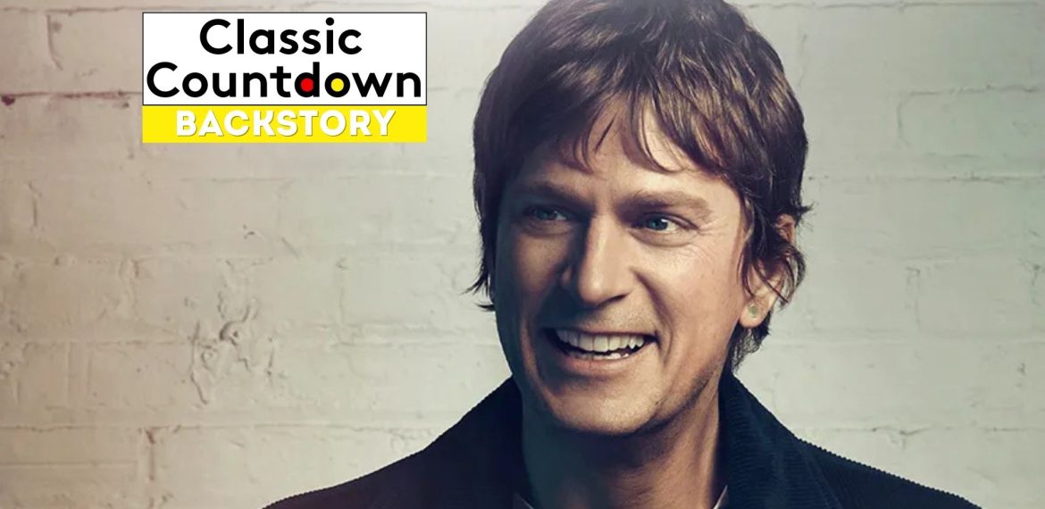 A smiling man with brown hair and a dark jacket stands in front of a light brick wall. In the upper left corner, a Classic Countdown Backstory logo appears, featuring bold black and yellow text inspired by "Last Christmas.