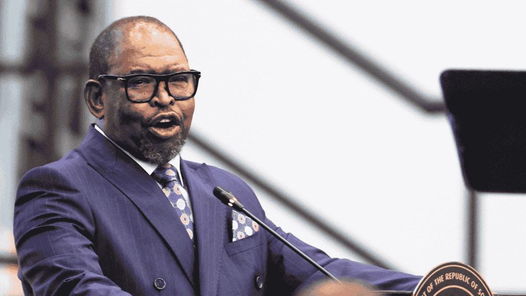 A man in a blue suit and tie speaks at a podium with a microphone, possibly addressing matric students. He is wearing glasses, and the blurred background draws full attention to the speaker.