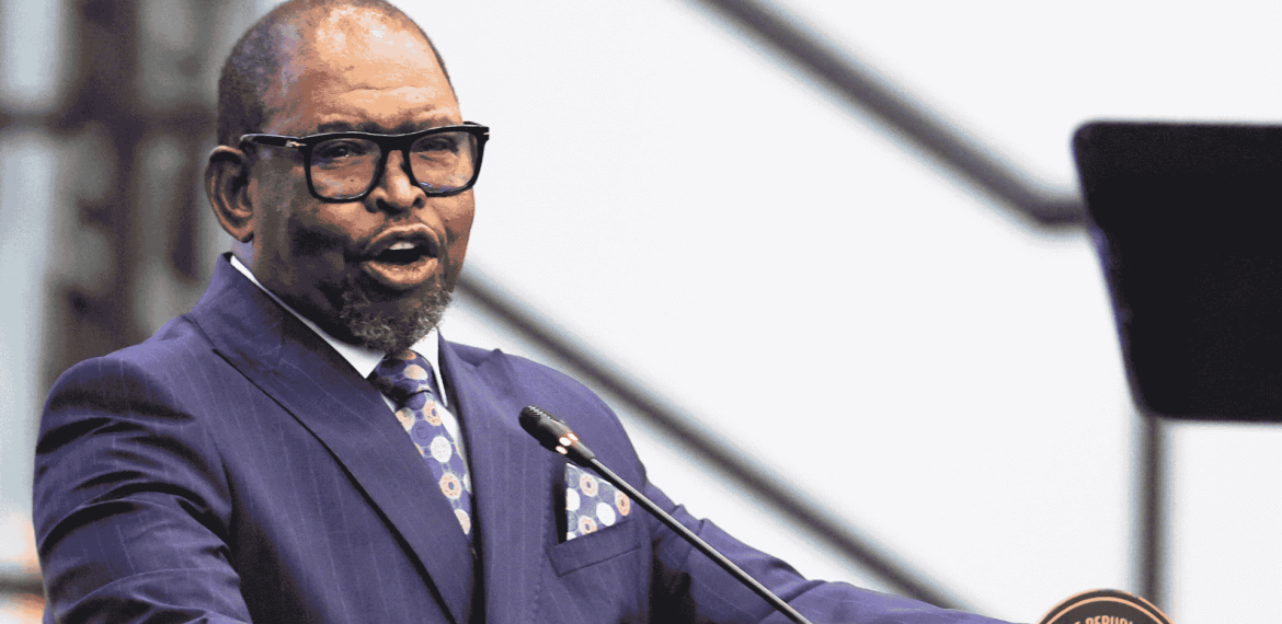 A man in a blue suit and tie speaks at a podium with a microphone, possibly addressing matric students. He is wearing glasses, and the blurred background draws full attention to the speaker.