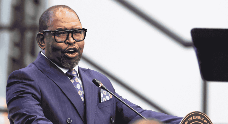 A man in a blue suit and tie speaks at a podium with a microphone, possibly addressing matric students. He is wearing glasses, and the blurred background draws full attention to the speaker.