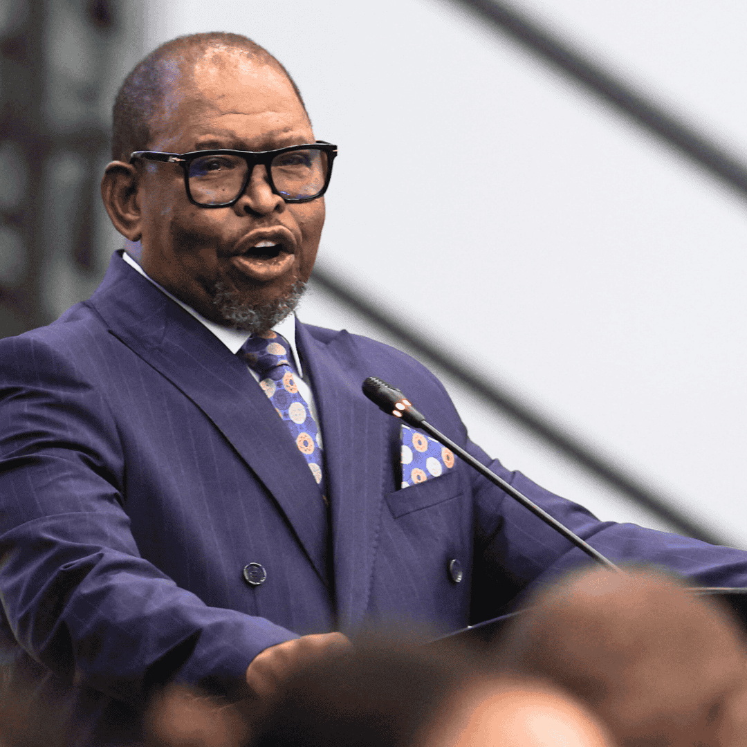 A man in a blue suit, patterned tie, and glasses speaks at a podium with a microphone, possibly addressing a Matric awards ceremony in an indoor setting.