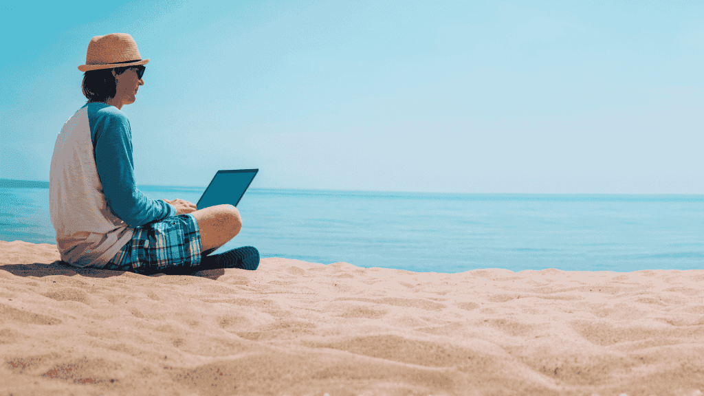 A person in a hat, sunglasses, and casual clothes sits on the sandy beach near the ocean, working on a Matric project on their laptop under a clear blue sky.