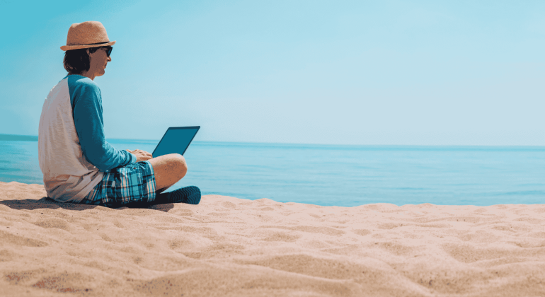 A person in a hat, sunglasses, and casual clothes sits on the sandy beach near the ocean, working on a Matric project on their laptop under a clear blue sky.
