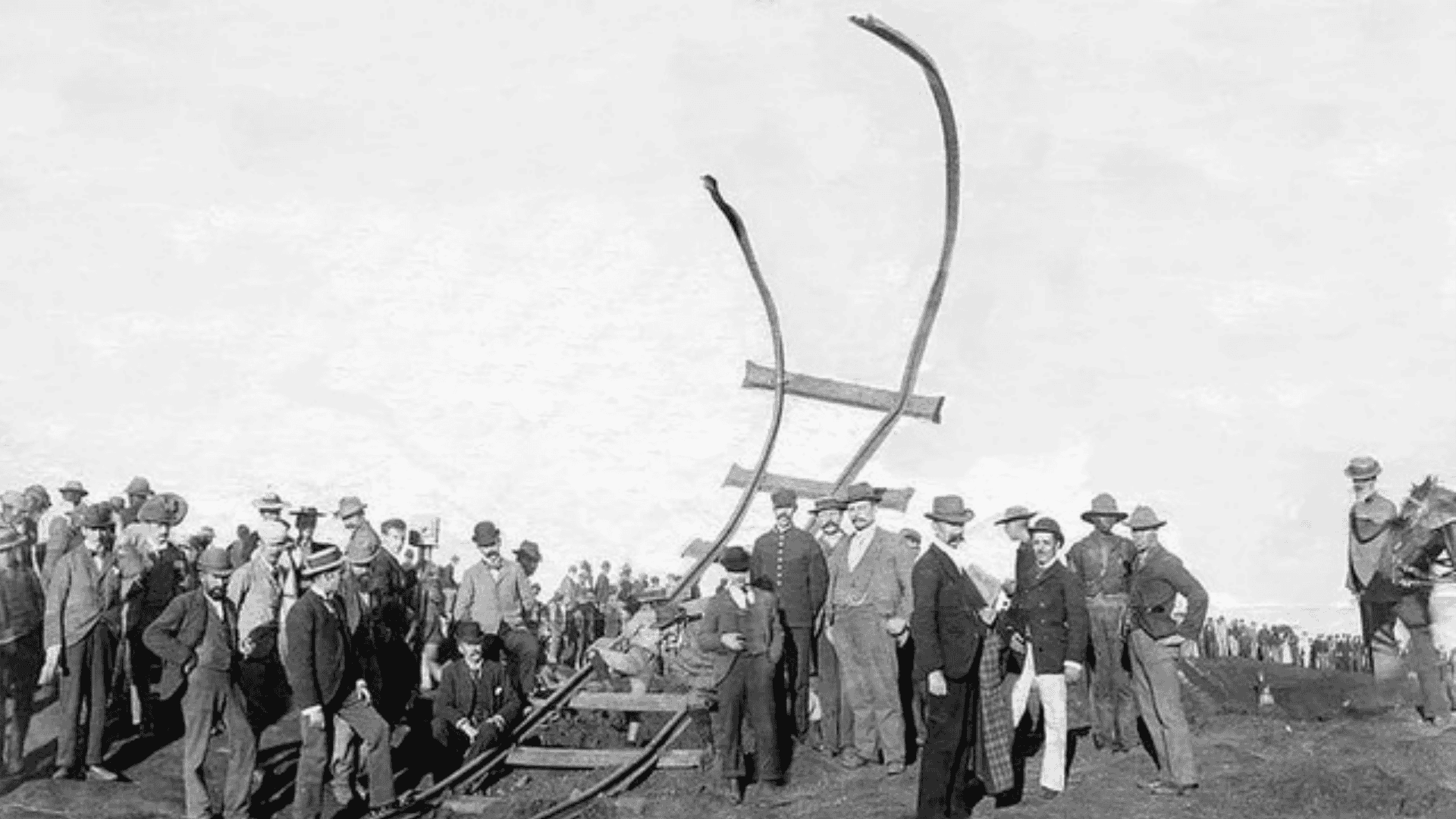 A large crowd of men in hats gathers around twisted, upturned railroad tracks on a barren landscape, suggesting an accident or disaster, while some people observe closely and others stand in groups, evoking a scene reminiscent of a Miriam Makeba song.