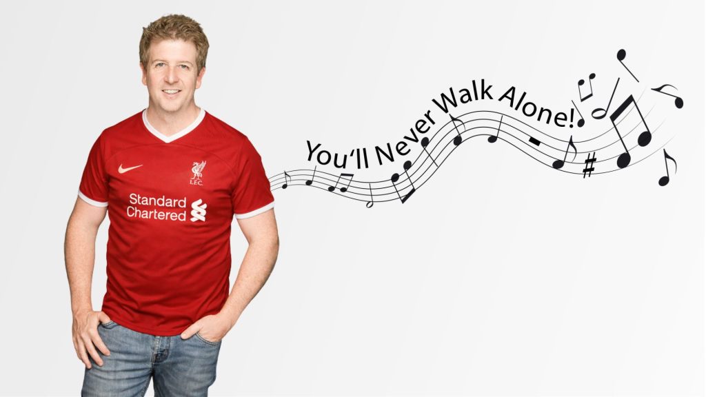 A smiling man in a red Liverpool football jersey stands beside musical notes and the text “You’ll Never Walk Alone!” flowing on a staff across a light background, echoing the uplifting vibe of Hot 1027 Breakfast.