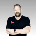 A bearded man with short hair smiles confidently with arms crossed, wearing a black HOT 102.7FM t-shirt and a smartwatch. Ian F stands against a light gray gradient background.