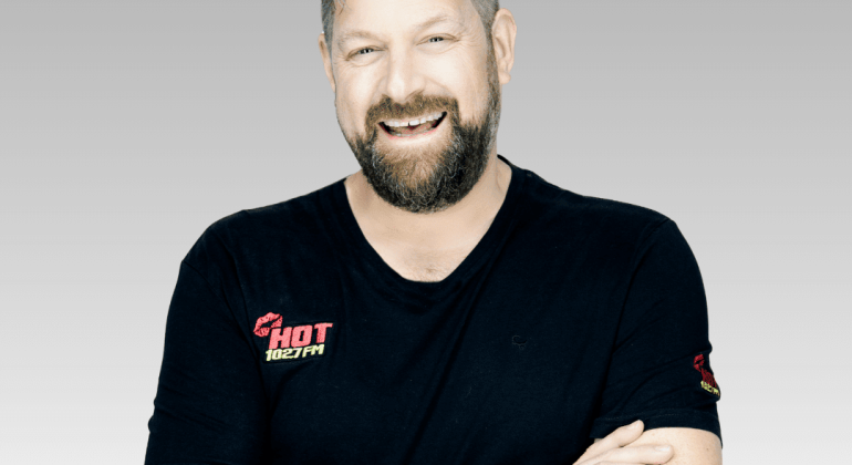 A bearded man with short hair smiles confidently with arms crossed, wearing a black HOT 102.7FM t-shirt and a smartwatch. Ian F stands against a light gray gradient background.