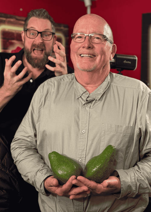 Two smiling men pose indoors; Ian F holds two large avocados in front, while the man behind him gestures with excitement, mouth open and hands raised. The red background features framed art and studio equipment.