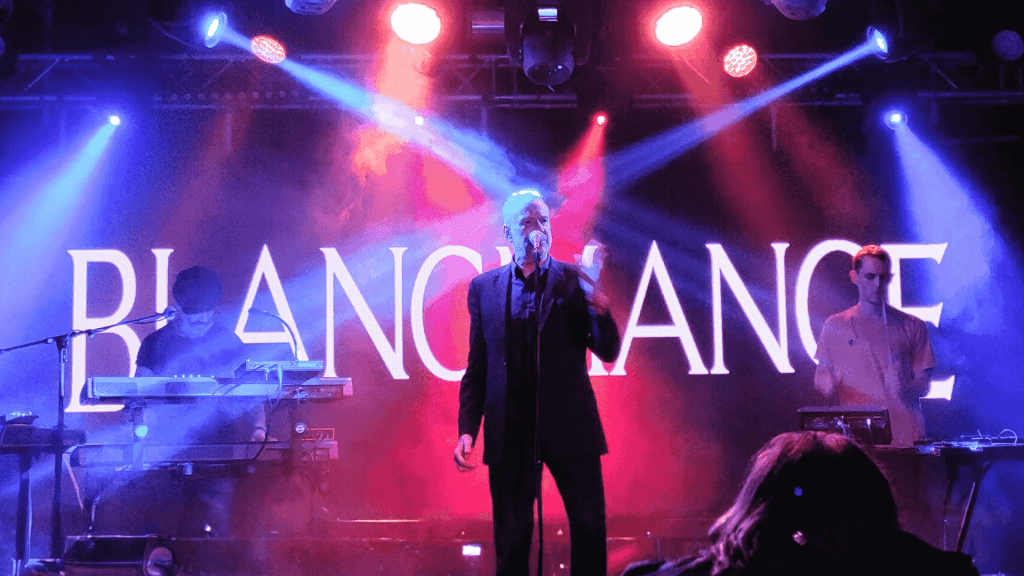 A band performs on a stage with dramatic blue and purple lighting. The lead singer stands at the center with a microphone, flanked by two musicians on keyboards. “BLANCMANGE” is displayed in large letters, reminiscent of a hot 1027 breakfast show backdrop.