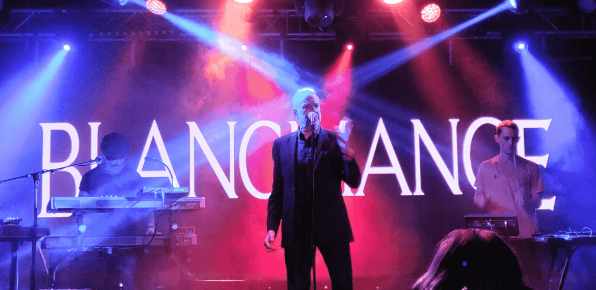 A band performs on a stage with dramatic blue and purple lighting. The lead singer stands at the center with a microphone, flanked by two musicians on keyboards. “BLANCMANGE” is displayed in large letters, reminiscent of a hot 1027 breakfast show backdrop.