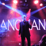 A band performs on a stage with dramatic blue and purple lighting. The lead singer stands at the center with a microphone, flanked by two musicians on keyboards. “BLANCMANGE” is displayed in large letters, reminiscent of a hot 1027 breakfast show backdrop.