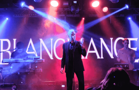 A band performs on a stage with dramatic blue and purple lighting. The lead singer stands at the center with a microphone, flanked by two musicians on keyboards. “BLANCMANGE” is displayed in large letters, reminiscent of a hot 1027 breakfast show backdrop.